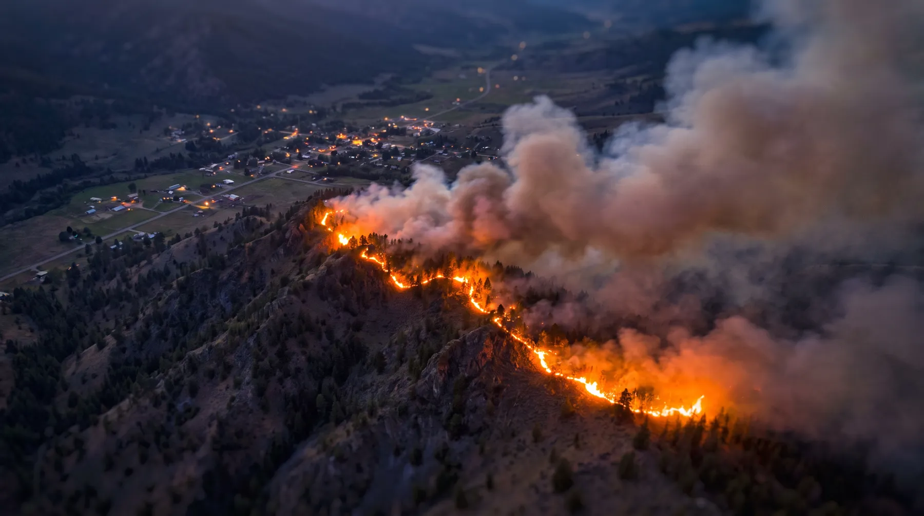 Wildfire burning along a forested ridge above a valley town at dusk